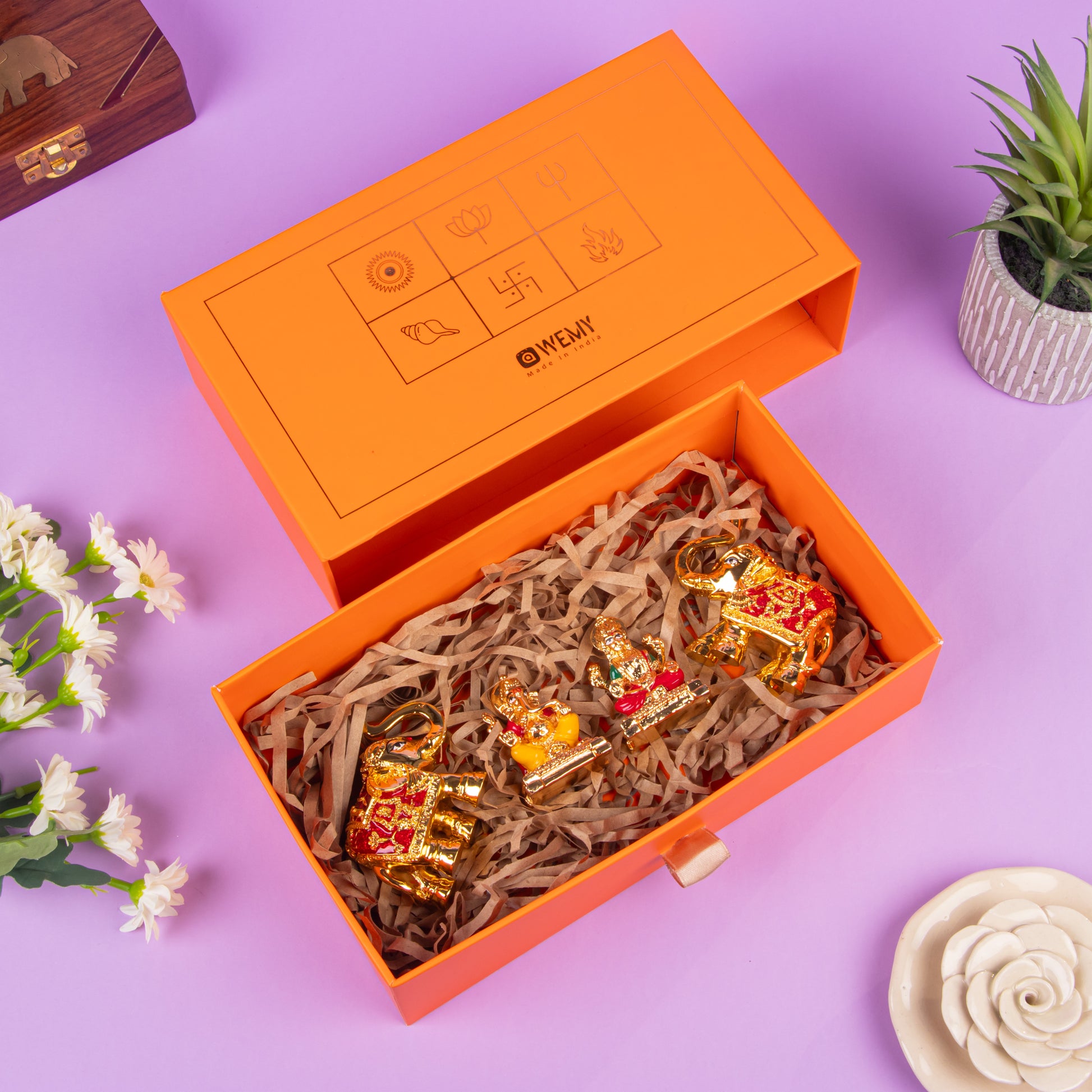 Elegant orange gift box with gold-plated decorative figurines of two elephants and a deity, presented on shredded paper, surrounded by flowers and plants on a purple background.