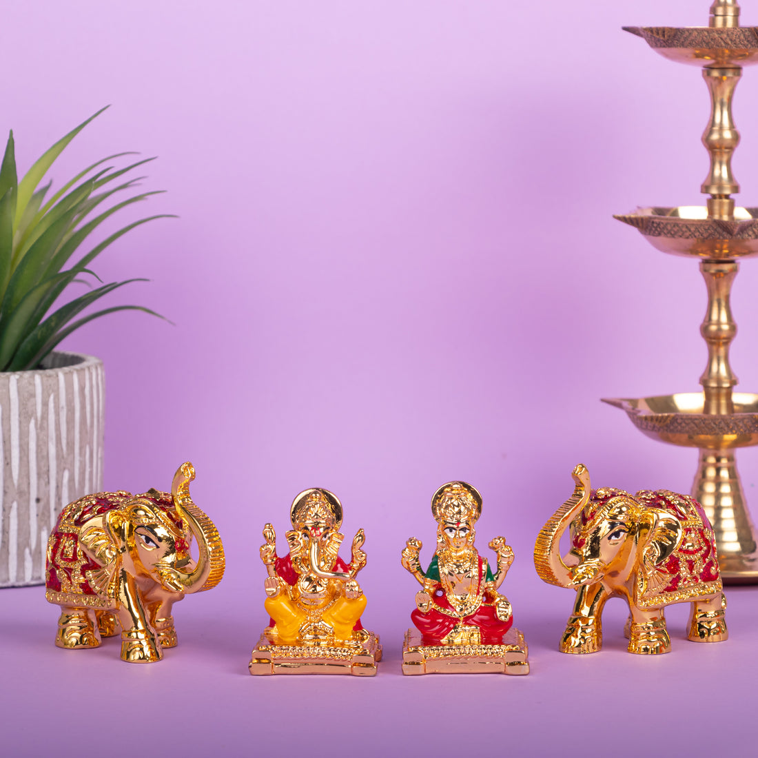 Gold and red idols of Lord Ganesha and Goddess Lakshmi sit centrally on a purple surface, flanked by two ornate elephant statues, with a potted plant on the left and a traditional tiered brass lamp on the right, against a light purple background.