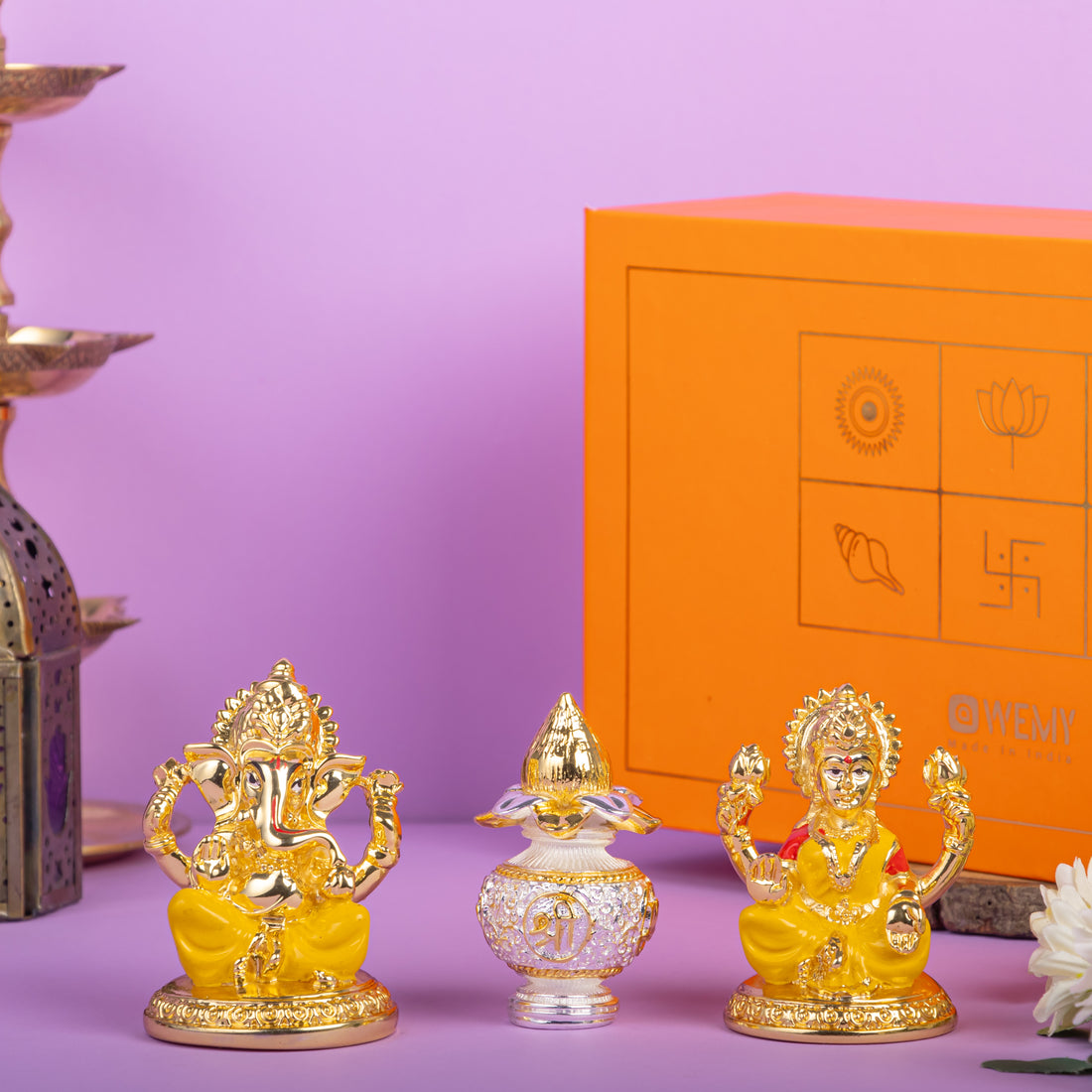 Golden idols of Lord Ganesha and Goddess Lakshmi with a decorative crystal kalash in between, placed in front of an orange gift box with traditional Indian symbols, on a purple background.