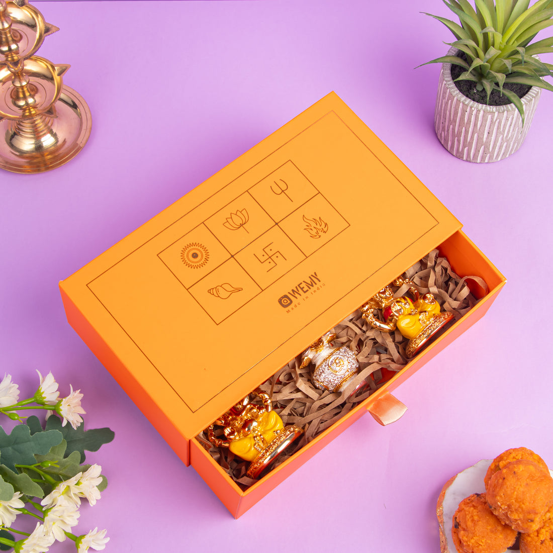Golden idols of Lord Ganesha and Goddess Lakshmi with a decorative crystal kalash in between, placed in front of an orange gift box with traditional Indian symbols, on a purple background.