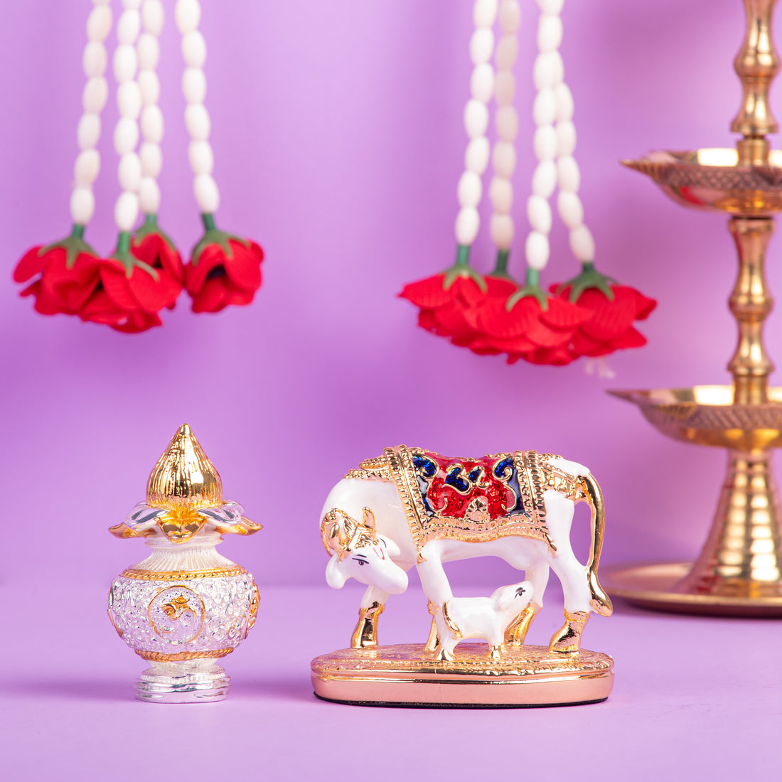 A still life arrangement featuring a white and gold Kamadhenu cow and calf idol with colorful accents, placed on a gold stand. To its left, a decorative gold and clear glass perfume bottle sits in front of an orange box with 'WEMY' written on it. In the background, white and red flower garlands hang, and a traditional Indian oil lamp (diya) is visible on the right.