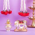 A beautiful, gold-finished Kamadhenu cow with calf idol, intricately detailed with red and blue accents, placed next to a silver-colored kalash and white floral garlands with red roses, all against a purple background.