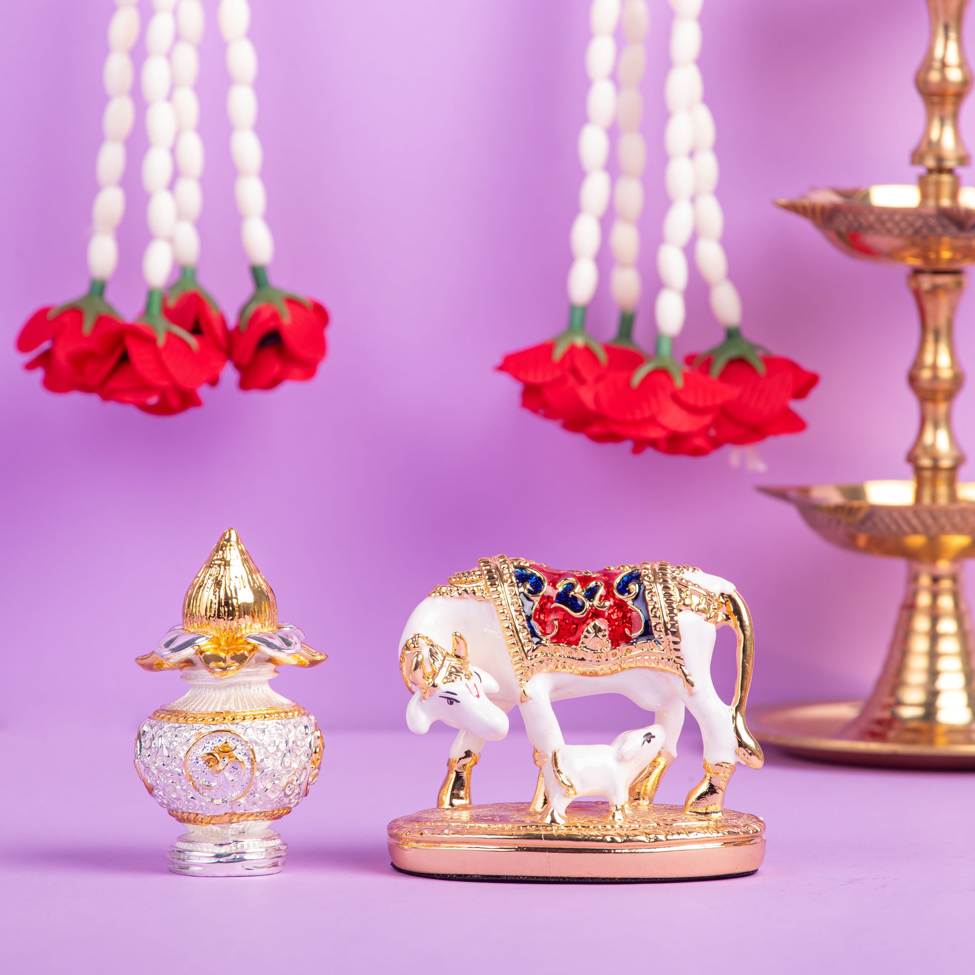 A beautiful, gold-finished Kamadhenu cow with calf idol, intricately detailed with red and blue accents, placed next to a silver-colored kalash and white floral garlands with red roses, all against a purple background.