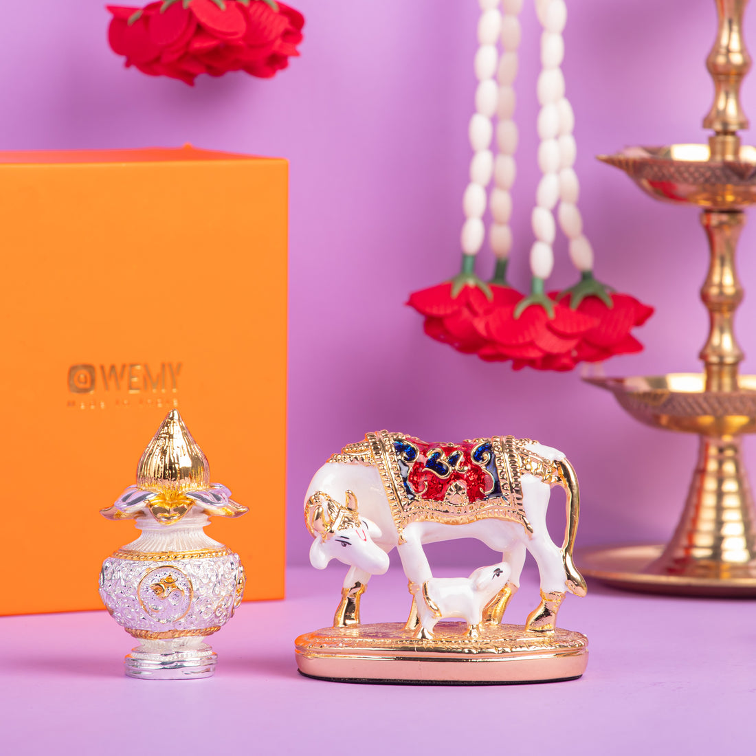 A still life arrangement featuring a white and gold Kamadhenu cow and calf idol with colorful accents, placed on a gold stand. To its left, a decorative gold and clear glass perfume bottle sits in front of an orange box with 'WEMY' written on it. In the background, white and red flower garlands hang, and a traditional Indian oil lamp (diya) is visible on the right.