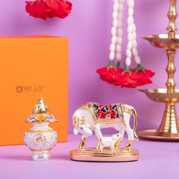 A still life arrangement featuring a white and gold Kamadhenu cow and calf idol with colorful accents, placed on a gold stand. To its left, a decorative gold and clear glass perfume bottle sits in front of an orange box with 'WEMY' written on it. In the background, white and red flower garlands hang, and a traditional Indian oil lamp (diya) is visible on the right.