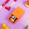 An image of an open orange gift box by WEMY, revealing two silver elephant idols and a silver kalash inside, nestled on a bed of shredded decorative material. The box sits on a purple surface, with floral garlands and other festive decor in the background, suggesting a gifting or festive occasion.