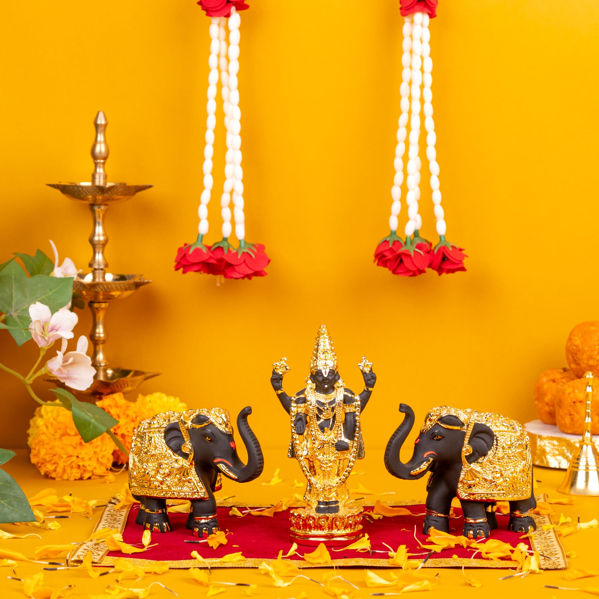 A black and gold Balaji idol flanked by two black and gold elephant statues, arranged on a red and gold mat with scattered yellow flower petals, against a vibrant yellow background with traditional Indian oil lamps and white floral garlands hanging above.