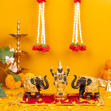 A black and gold Balaji idol flanked by two black and gold elephant statues, arranged on a red and gold mat with scattered yellow flower petals, against a vibrant yellow background with traditional Indian oil lamps and white floral garlands hanging above.