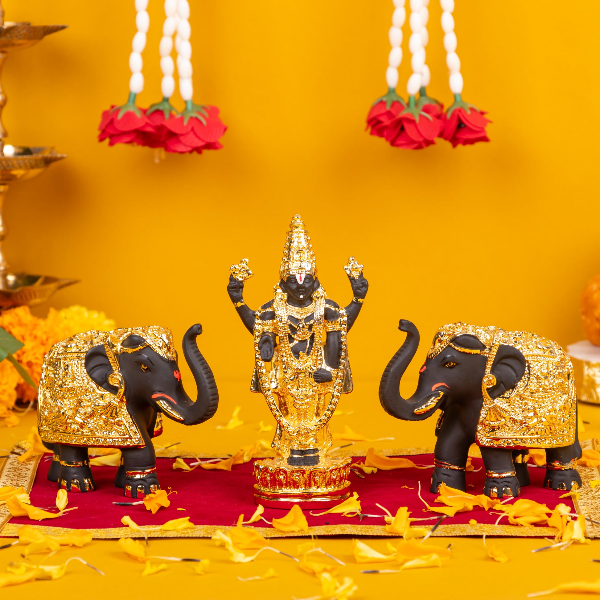 A black and gold Lord Balaji idol flanked by two black and gold elephant statues, arranged on a red cloth adorned with scattered yellow and orange flower petals, with red and white floral garlands hanging in the golden-yellow background.