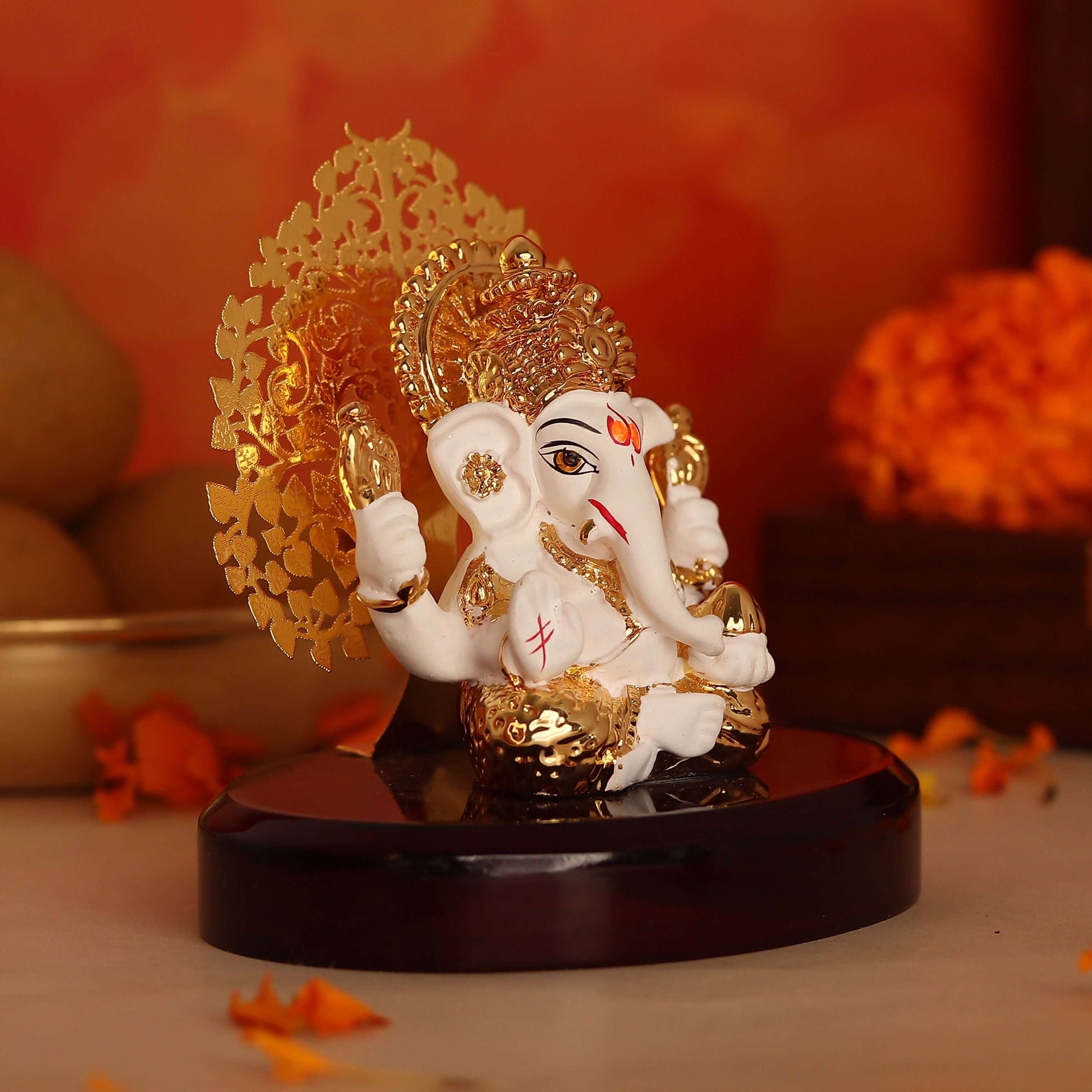 A white and gold Ganesha idol with a golden floral backdrop on a dark wooden base.