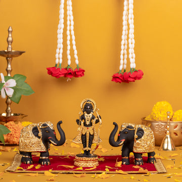 A Hindu deity idol, likely Ram Lalla, flanked by two golden-adorned elephant statues, set up for worship on a red cloth with garlands and a traditional oil lamp in the background.