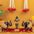 A detailed image of a black Ram Lalla idol adorned with gold ornaments, standing on a lotus base on a red cloth scattered with yellow flower petals, flanked by two black and gold elephant statues with trunks raised, with white and red floral garlands hanging in the background.