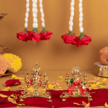 A pair of small golden idols, featuring Lord Ganesha on the left and Goddess Lakshmi on the right, adorned with red and white floral garlands, set against a warm-toned background with scattered flower petals.