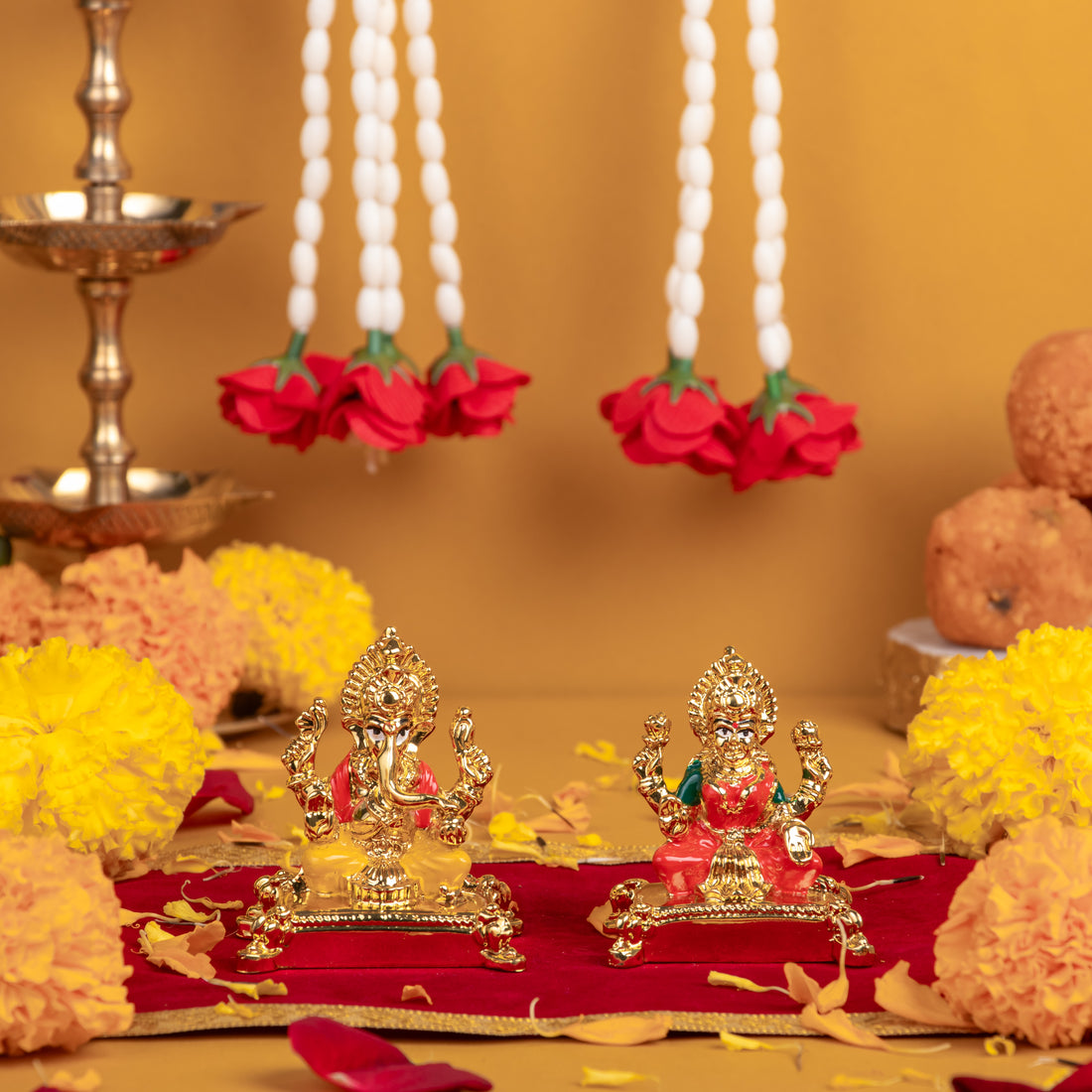 A pair of small golden idols, featuring Lord Ganesha on the left and Goddess Lakshmi on the right, adorned with red and white floral garlands, set against a warm-toned background with scattered flower petals.
