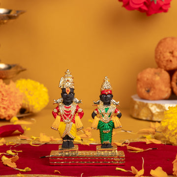 Two intricately crafted statues of Hindu deities, Lord Vitthal and Goddess Rukmini, standing on a gilded base on a red velvet cloth. Lord Vitthal, on the left, wears a yellow dhoti and green shawl, while Rukmini, on the right, wears a green saree. Both are adorned with elaborate gold crowns, necklaces, and other jewelry, with a warm-toned background featuring diffused flowers and offerings