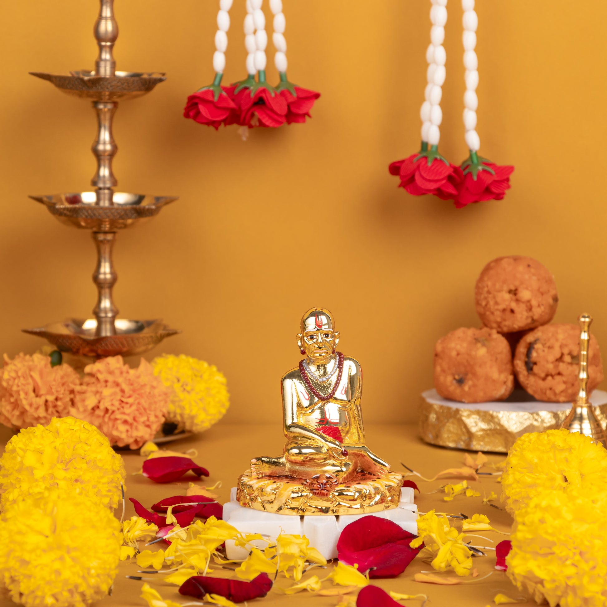 A gold-colored Swami Samarth idol seated on a base, surrounded by yellow and red flowers, brass peacock diyas, and laddoos, set against a warm yellow background.