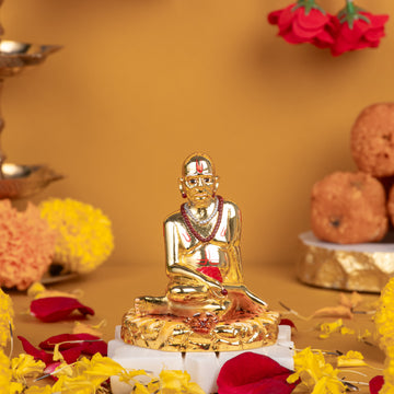A close-up of a golden Swami Samarth idol seated in a meditative pose, adorned with a red tilak and garland, surrounded by traditional offerings like marigold petals, red rose petals, and ladoos, against a warm yellow background.