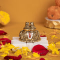 A close-up of a gold-plated Lord Balaji Charan Paduka (divine footprints) with an antique finish, intricately detailed with sacred symbols like the Shanku (conch), Chakra (disc), and Namam (U-shaped mark), resting on a white pedestal amidst scattered red rose petals and yellow marigold flowers.