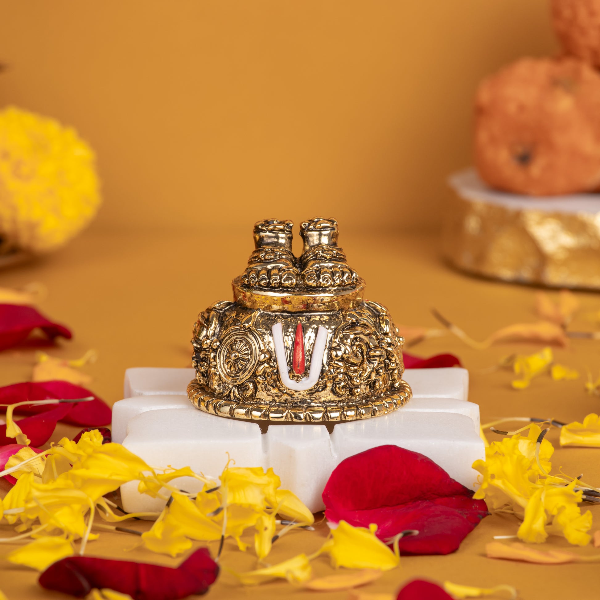 A close-up of a gold-plated Lord Balaji Charan Paduka (divine footprints) with an antique finish, intricately detailed with sacred symbols like the Shanku (conch), Chakra (disc), and Namam (U-shaped mark), resting on a white pedestal amidst scattered red rose petals and yellow marigold flowers.
