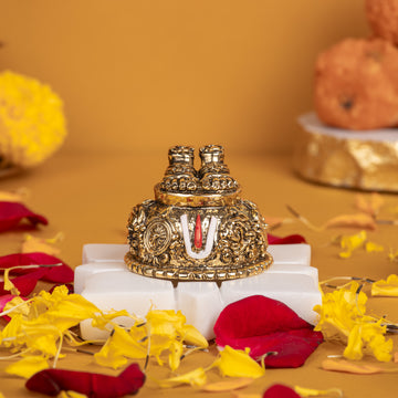 A close-up of a gold-plated Lord Balaji Charan Paduka (divine footprints) with an antique finish, intricately detailed with sacred symbols like the Shanku (conch), Chakra (disc), and Namam (U-shaped mark), resting on a white pedestal amidst scattered red rose petals and yellow marigold flowers.
