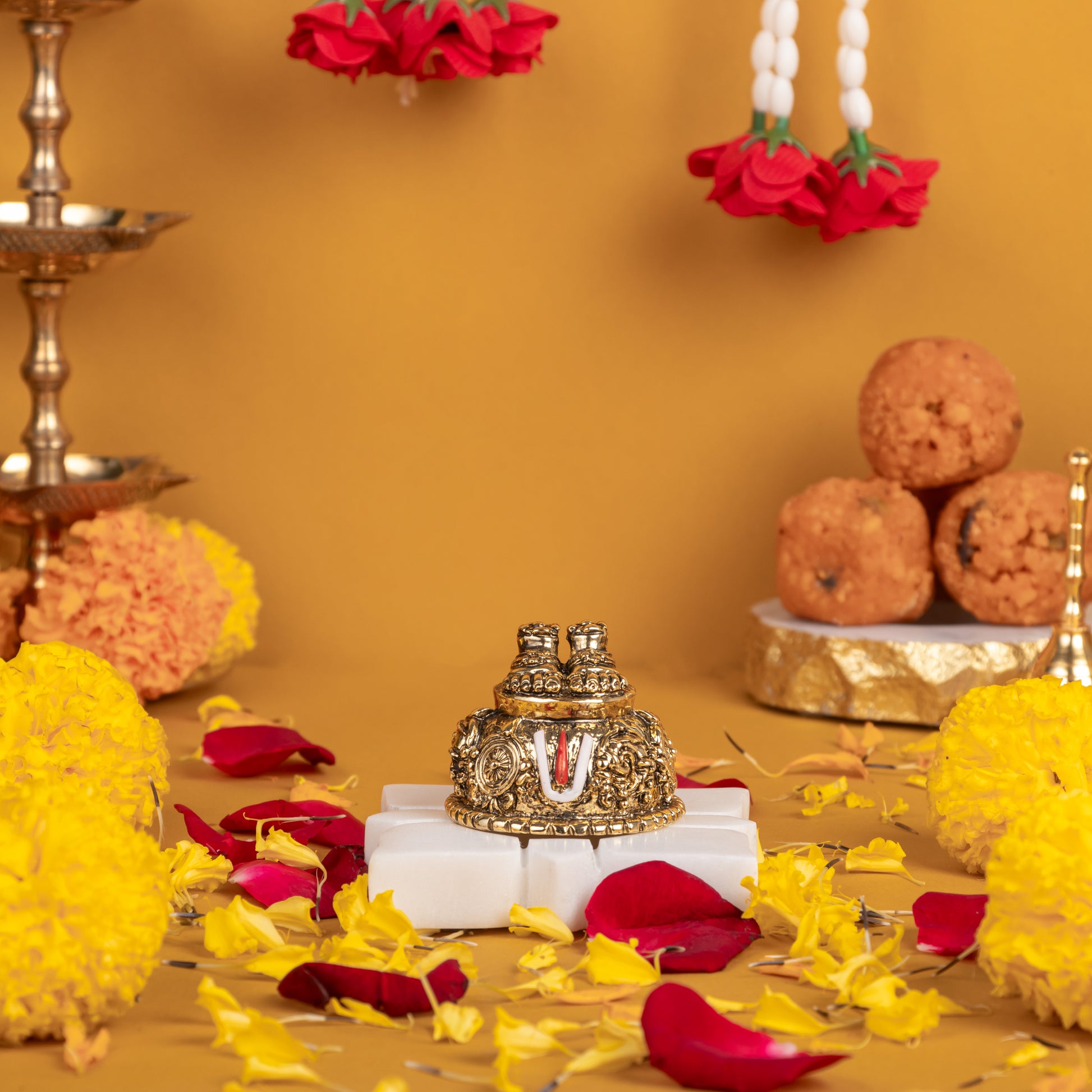 A golden Balaji Charan Paduka (divine feet) on a white stand, surrounded by yellow marigolds, scattered red rose petals, a traditional brass oil lamp, and three ladoo sweets, all set against a warm golden-orange background.