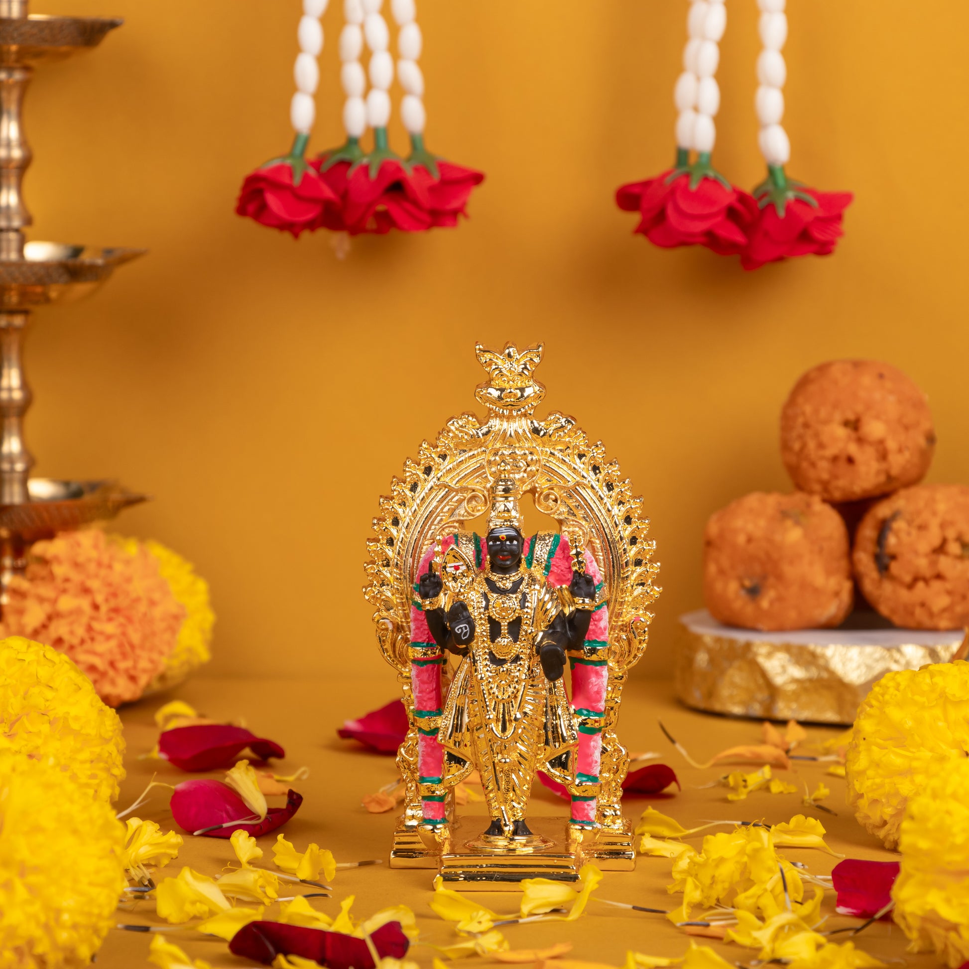 Golden colored idol of Lord Murugan (Kartikeya/Subrahmanya) standing on a decorative base, adorned with pink and green accents, surrounded by offerings of yellow marigold flowers, red rose petals, pearl garlands with red roses, and a stack of Indian sweet laddus on a gold background.