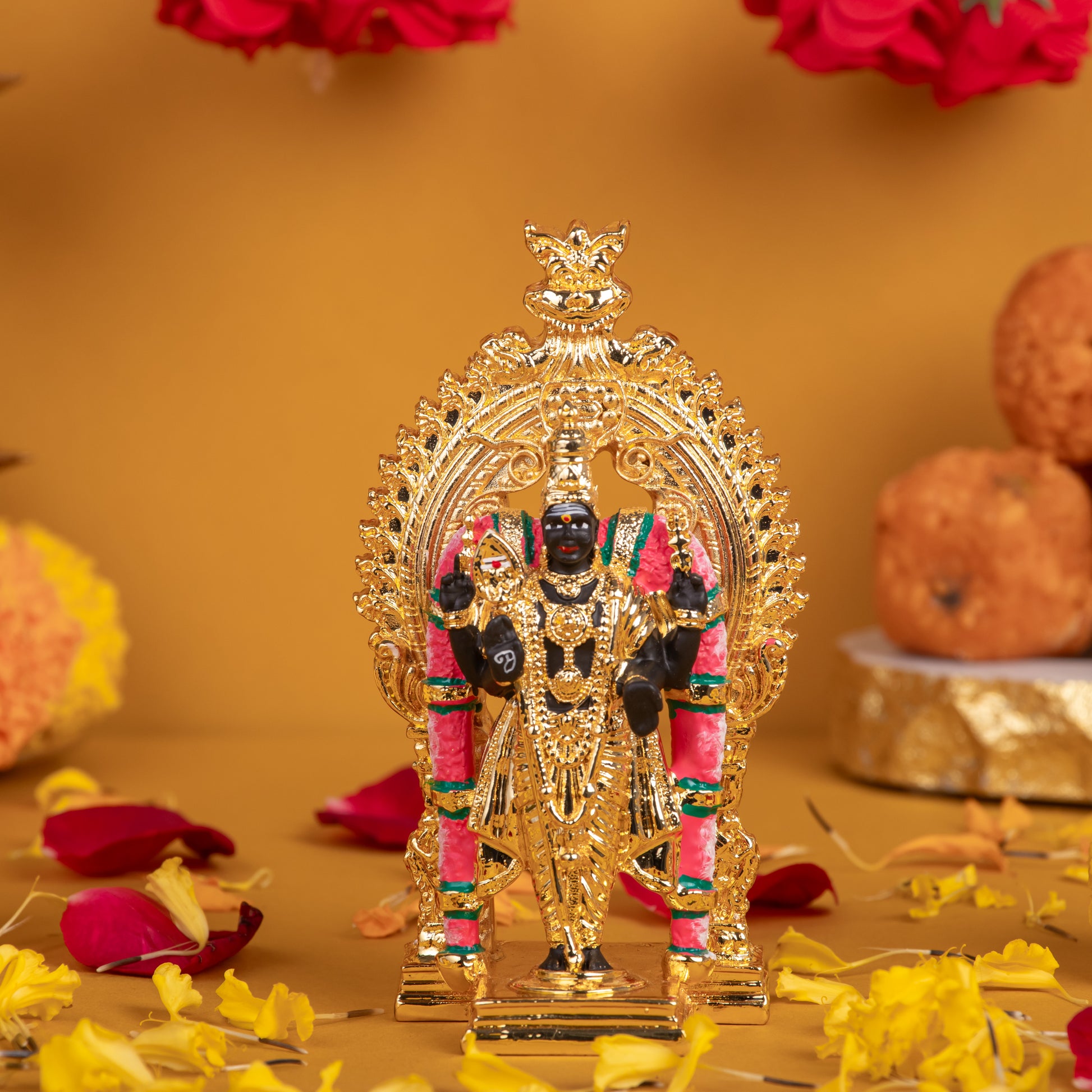 A detailed, golden idol of Lord Murugan, standing upright on a decorative base, adorned with intricate carvings and colorful accents, set against a warm yellow-orange background with scattered red and yellow flower petals.