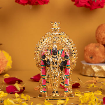 A detailed, golden idol of Lord Murugan, standing upright on a decorative base, adorned with intricate carvings and colorful accents, set against a warm yellow-orange background with scattered red and yellow flower petals.