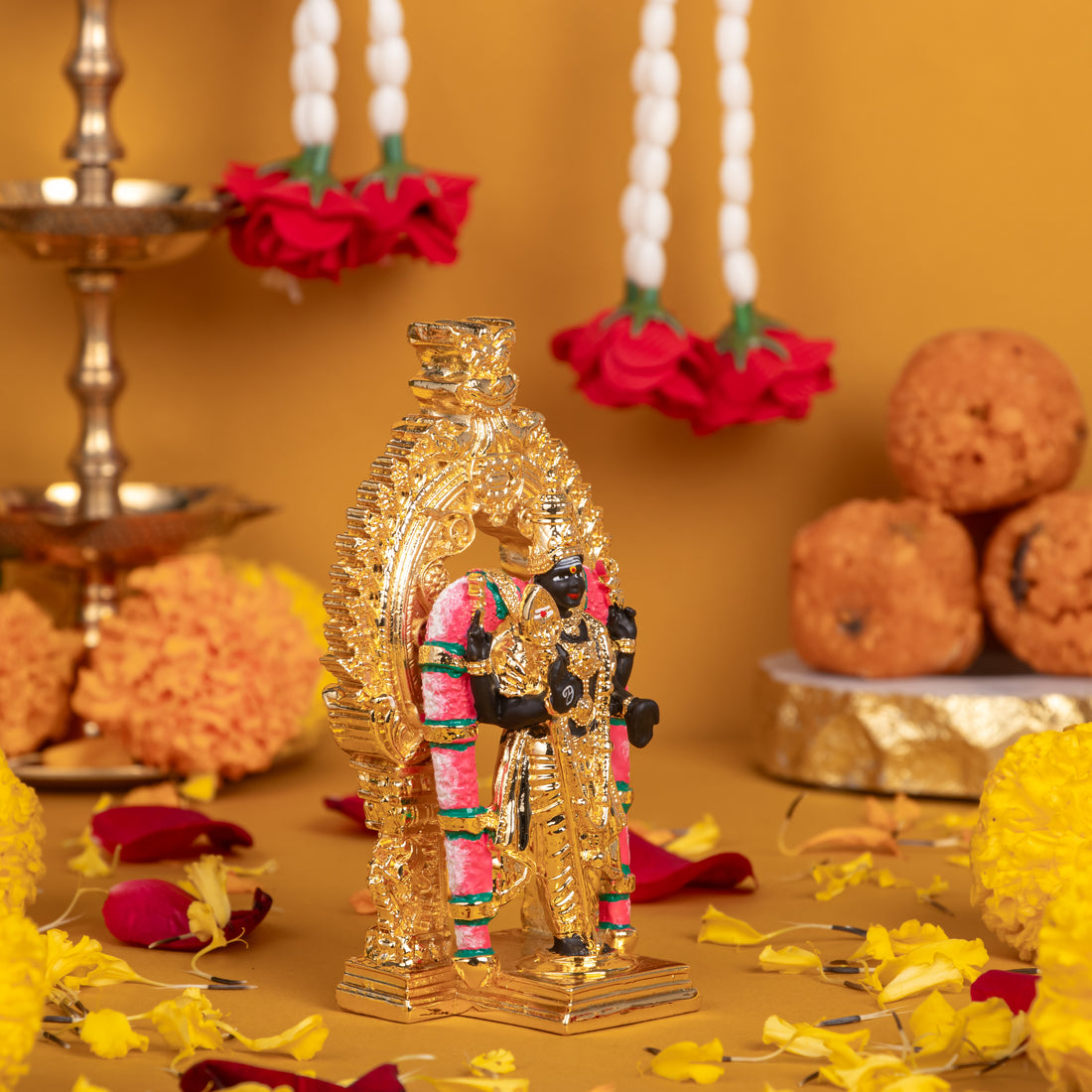 Golden colored idol of Lord Murugan (Kartikeya/Subrahmanya) standing on a decorative base, adorned with pink and green accents, surrounded by offerings of yellow marigold flowers, red rose petals, pearl garlands with red roses, and a stack of Indian sweet laddus on a gold background.