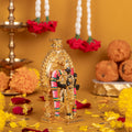 A vibrant gold-plated idol of Lord Murugan, adorned with a colorful garment, stands amidst scattered yellow marigold petals. In the background, white and red flower garlands hang, and a traditional oil lamp along with a stack of ladoo sweets are visible, all set against a warm yellow backdrop
