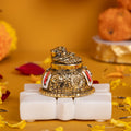A detailed image of a gold-plated Lord Balaji Charan Paduka (footprints of Lord Venkateswara), featuring the sacred Shanku (conch), Chakra (discus), and Namam symbols on a white stand, against a warm yellow background with scattered flower petals.