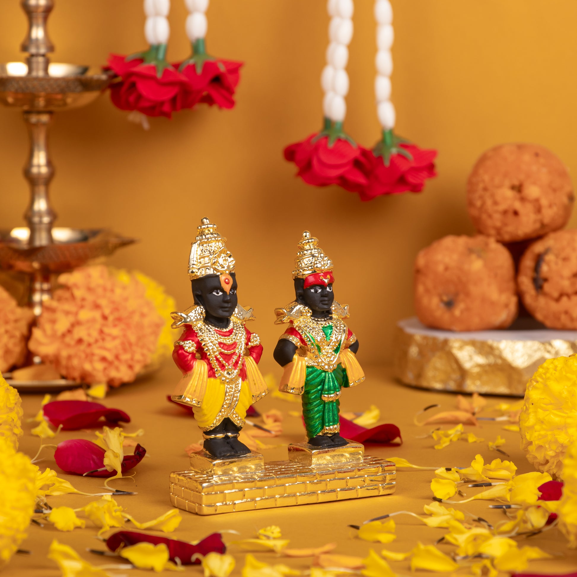 Ornate idols of Lord Vitthal and Goddess Rukmini in traditional attire, adorned with fresh flower garlands, yellow marigolds, scattered rose petals, and ladoo offerings on a vibrant yellow background, capturing a sacred festive scene.