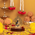 A black and gold Lord Buddha statue is seated on a dark wooden base. An intricate golden halo surrounds the statue. The scene is decorated with yellow and orange flowers, red and yellow petals, and two hanging floral garlands with red flowers. Traditional sweets and a lamp are also visible in the background.