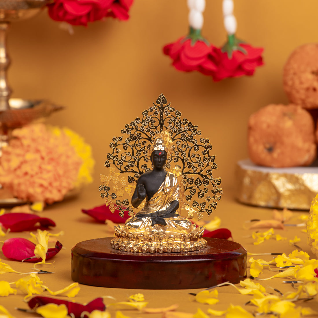 A golden and black Lord Buddha Statue seated on a wooden base with a golden, intricate Bodhi tree background. The idol is surrounded by a floral arrangement with yellow and red petals scattered on a yellow surface.