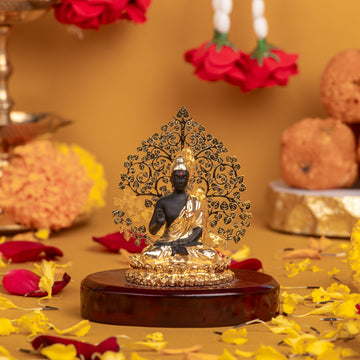 A golden and black Lord Buddha Statue seated on a wooden base with a golden, intricate Bodhi tree background. The idol is surrounded by a floral arrangement with yellow and red petals scattered on a yellow surface.