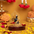 A golden and black Lord Buddha statue is seated on a wooden base with a golden, intricate Bodhi tree background. The statue is surrounded by a floral arrangement with yellow and red petals scattered on a yellow surface.