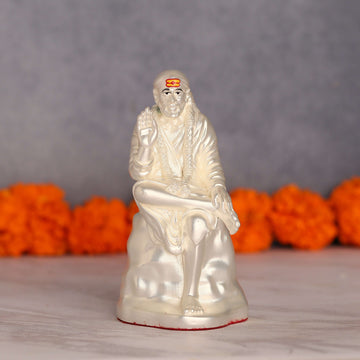 A white idol of Shree Sai Baba seated, with a prominent orange tilak on his forehead, and his right hand raised in a blessing gesture, set against a blurry grey background with orange marigold flowers.