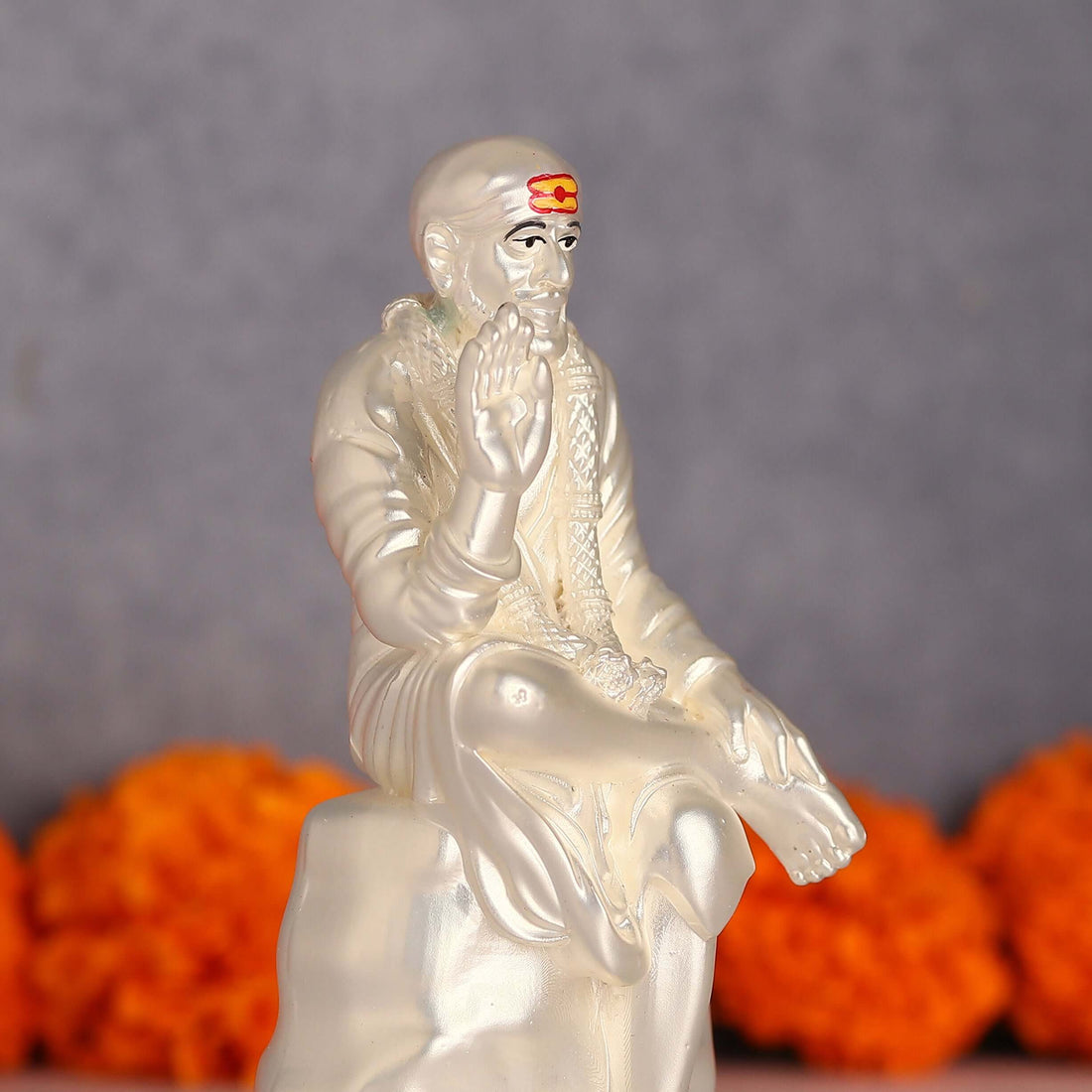 A white idol of Shree Sai Baba seated, with a prominent orange tilak on his forehead, and his right hand raised in a blessing gesture, set against a blurry grey background with orange marigold flowers.