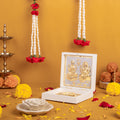 Golden idols of Goddess Lakshmi and Lord Ganesha seated on a lotus, housed in an open white decorative box, surrounded by traditional Hindu puja items like marigold and rose garlands, scattered flower petals, and a brass diya on a golden yellow background.
