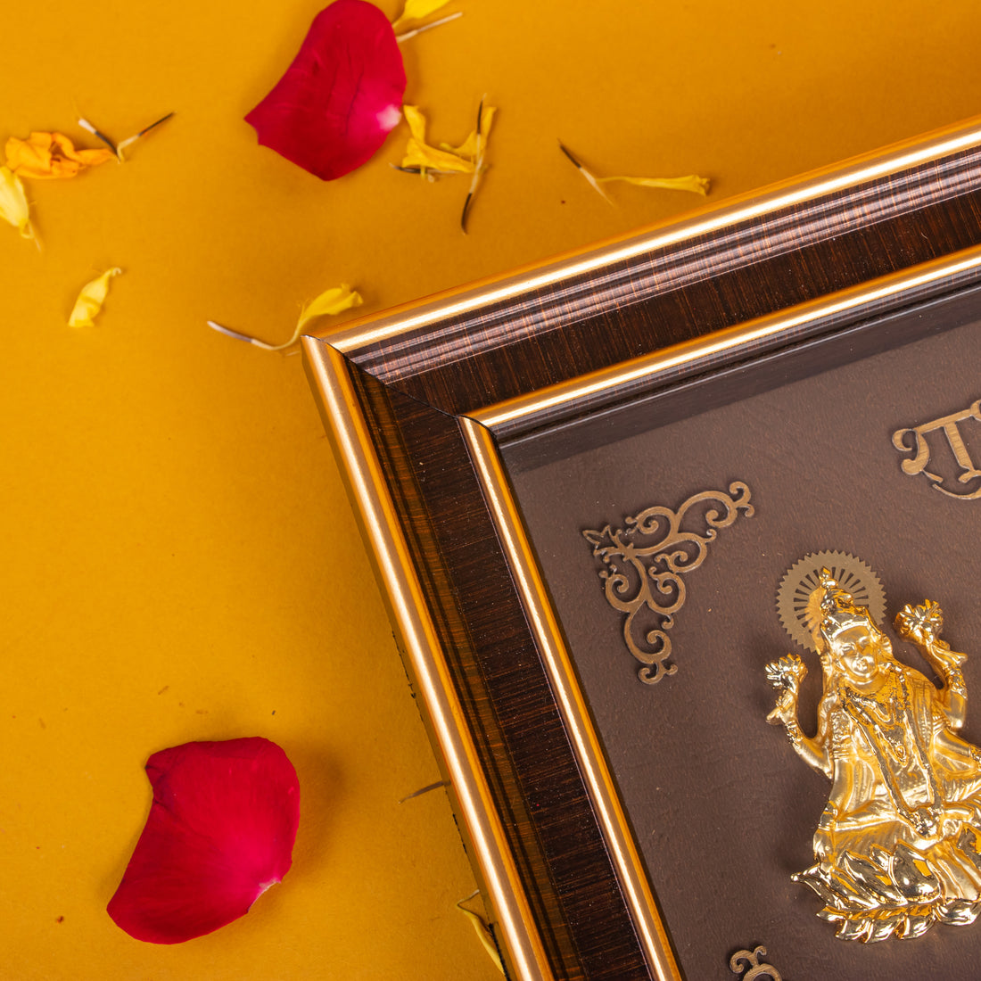 A brown framed photo with golden idols of Hindu deities Lakshmi, Ganesha, and Saraswati, labeled in Hindi script as "Shubh" (शुभ) and "Labh" (लाभ). The frame sits on a surface with scattered red flower petals and white flower garlands hanging in the background against a yellow wall.