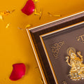 A brown and gold photo frame featuring golden idols of Hindu deities, likely the Trimurti, set against a dark background with intricate gold accents. The frame is positioned on a vibrant yellow surface scattered with loose red rose petals and a few yellow flower petals.