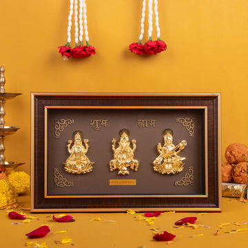 A brown framed photo with golden idols of Hindu deities Lakshmi, Ganesha, and Saraswati, labeled in Hindi script as "Shubh" (शुभ) and "Labh" (लाभ). The frame sits on a surface with scattered red flower petals and white flower garlands hanging in the background against a yellow wall.