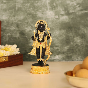 A black polyresin statue of Ram Lalla, Lord Rama as a young child, standing with a golden bow and arrow, adorned with gold ornaments, against a blurred green and brown background.