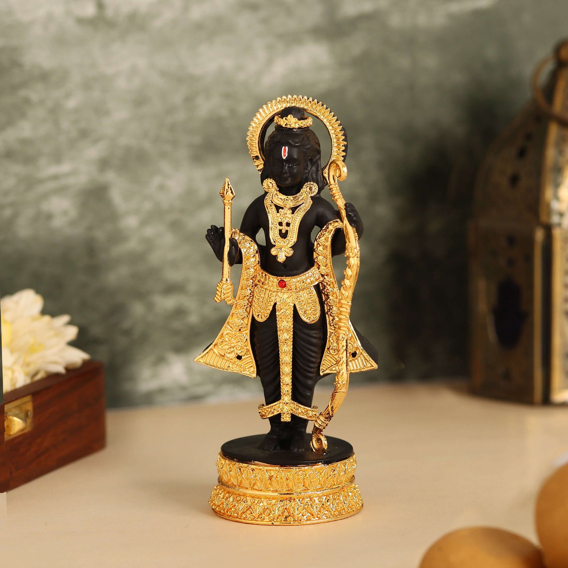 A striking black and gold statue of Lord Ram Lalla, adorned with traditional ornaments and holding a golden bow and arrow, stands on an ornate base with an elaborate halo behind his head.