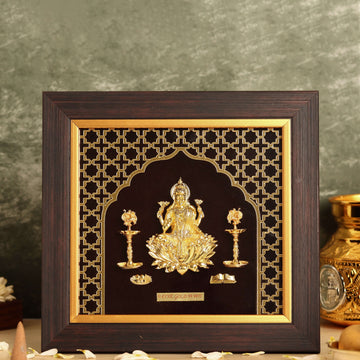 A photo frame with a dark wooden border features a golden relief of Goddess Laxmi, seated on a lotus flower. The intricate golden design is set against a black background with a geometric pattern. The goddess is flanked by two lit golden lamps, and the entire scene is enclosed in a golden archway.