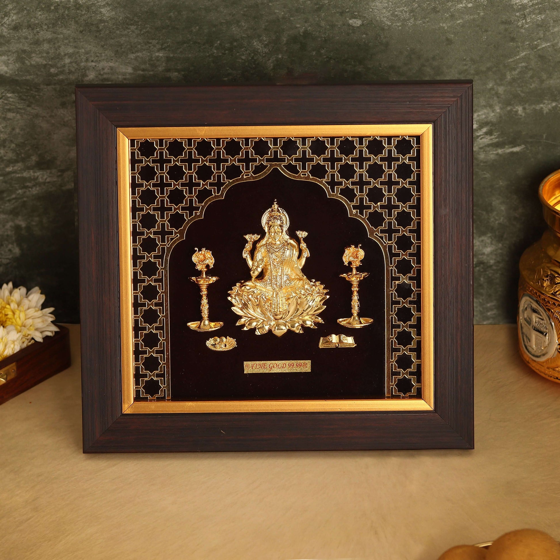 A photo frame with a dark wooden border features a golden relief of Goddess Laxmi, seated on a lotus flower. The intricate golden design is set against a black background with a geometric pattern. The goddess is flanked by two lit golden lamps, and the entire scene is enclosed in a golden archway.
