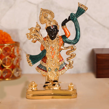 A meticulously crafted idol of Lord Shreenath Ji (a form of Lord Krishna) standing upright, adorned with intricate golden ornaments and vibrant green and orange garments, on a golden base. The idol features dark skin, a prominent nose, and large, expressive eyes, with one arm raised and the other holding a staff or a collection of flowers. The overall impression is one of divine beauty and reverence.