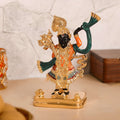 A vibrant, detailed idol of Lord Shreenath Ji, a child manifestation of Lord Krishna, stands prominently on a golden base. The deity has dark skin and is adorned with intricate gold jewelry, a large crown, and colorful garments primarily in orange and green, with subtle blue and red accents on the lower attire. One hand is raised, likely depicting the lifting of the Govardhan Hill, while the other rests at the waist in a fist.