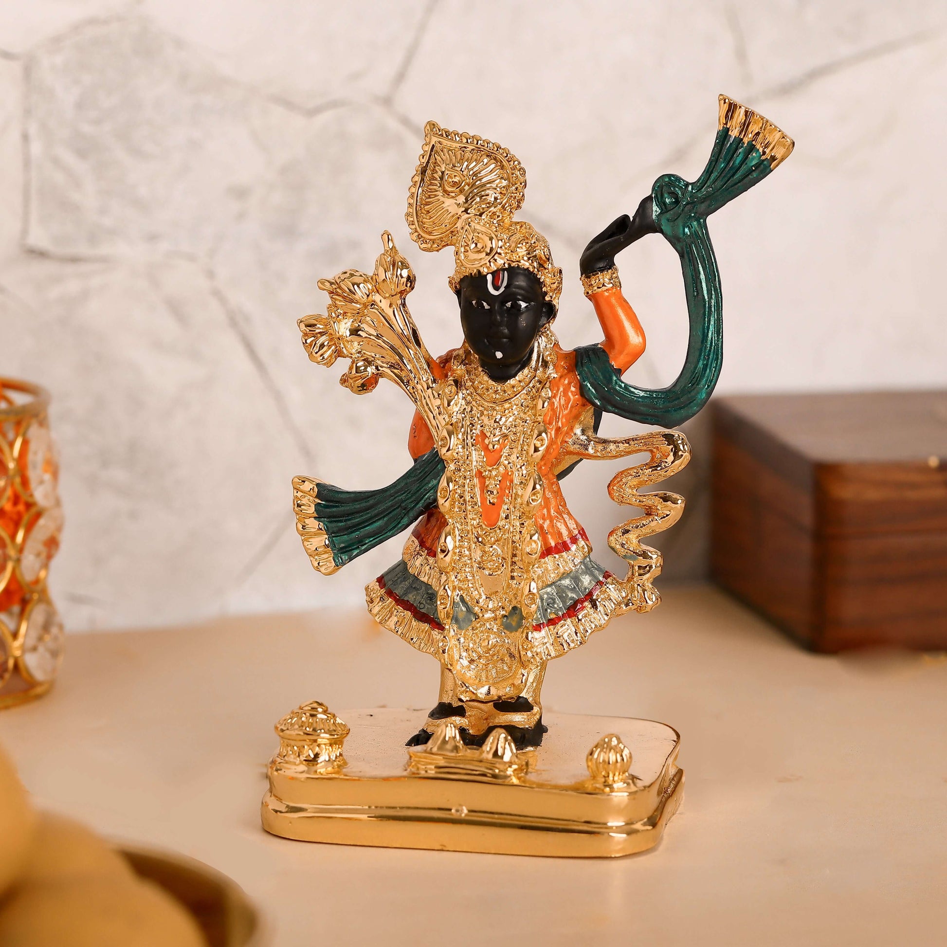 A vibrant, detailed idol of Lord Shreenath Ji, a child manifestation of Lord Krishna, stands prominently on a golden base. The deity has dark skin and is adorned with intricate gold jewelry, a large crown, and colorful garments primarily in orange and green, with subtle blue and red accents on the lower attire. One hand is raised, likely depicting the lifting of the Govardhan Hill, while the other rests at the waist in a fist.
