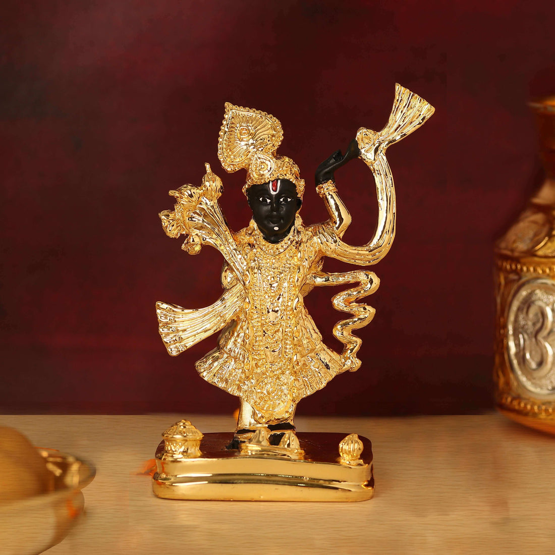 A meticulously crafted idol of Lord Shreenath Ji (a form of Lord Krishna) standing upright, adorned with intricate golden ornaments and vibrant green and orange garments, on a golden base. The idol features dark skin, a prominent nose, and large, expressive eyes, with one arm raised and the other holding a staff or a collection of flowers. The overall impression is one of divine beauty and reverence.